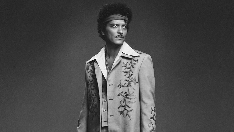 Black and white studio portrait of musician Bruno Mars wearing a headband and an embroidered 1970s-style suit coat.